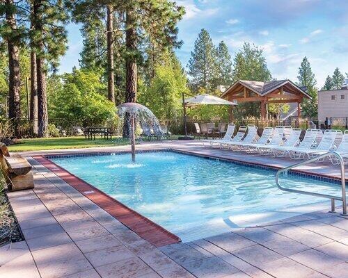 A relaxing outdoor pool area is surrounded by tall trees, featuring a fountain in the center. Sun loungers are positioned around the pool, with a shaded pavilion visible in the background, offering a space for gathering.