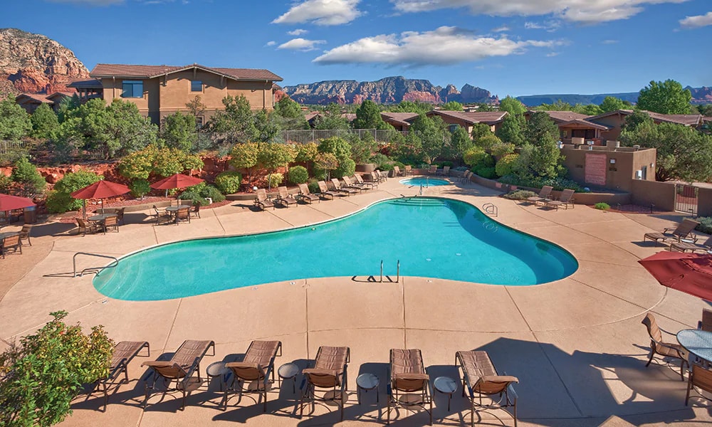 An outdoor pool is framed by lounge chairs and umbrellas, situated amidst landscaped greenery. The pool features a heart shape, with clear blue water under a blue sky, while a scenic backdrop of red rock formations can be seen in the distance.