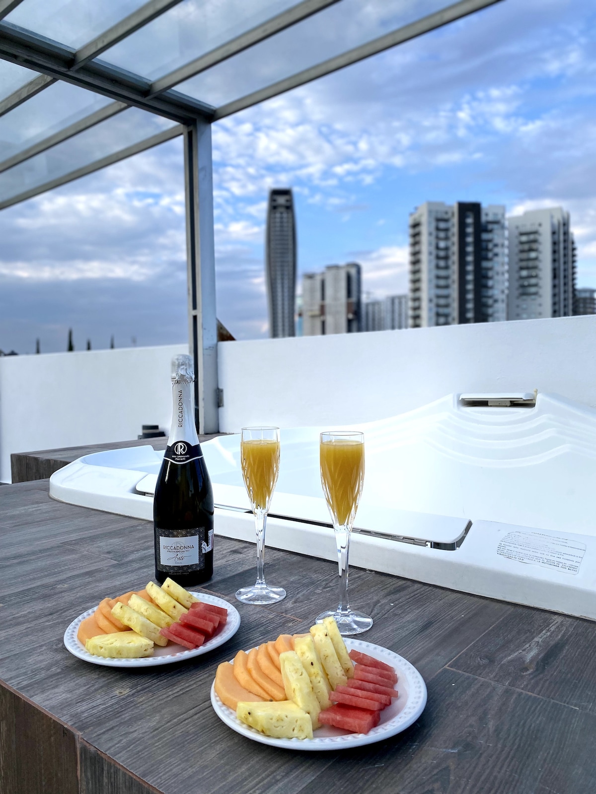 A rooftop space features a jacuzzi alongside a wooden table set with two plates of fresh fruit, including watermelon, cantaloupe, and pineapple. Two flutes filled with a sparkling beverage sit beside a bottle of champagne. A city skyline with tall buildings is visible in the background.