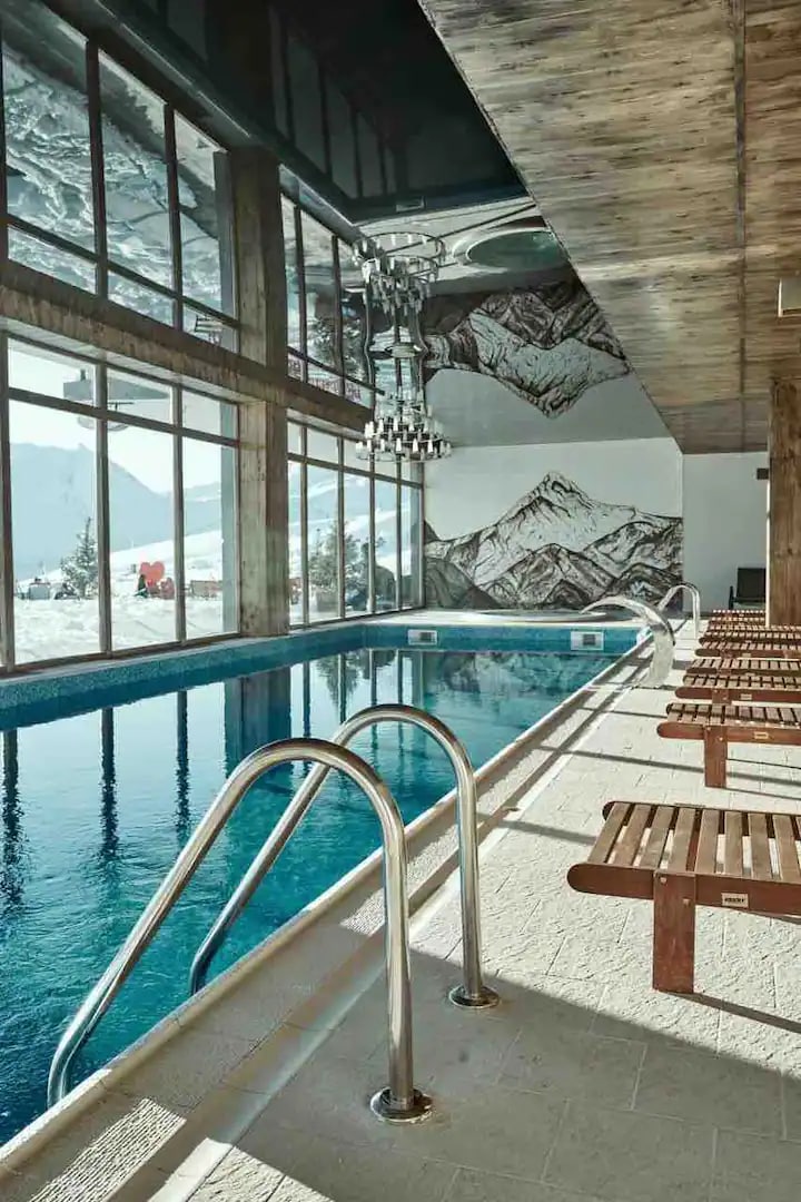 An indoor pool area showcases a beautifully maintained pool with clear blue water. Sunlight filters through expansive windows, illuminating the space and highlighting a stunning mountain mural on the wall. Wooden lounge chairs are arranged along the edge of the pool.