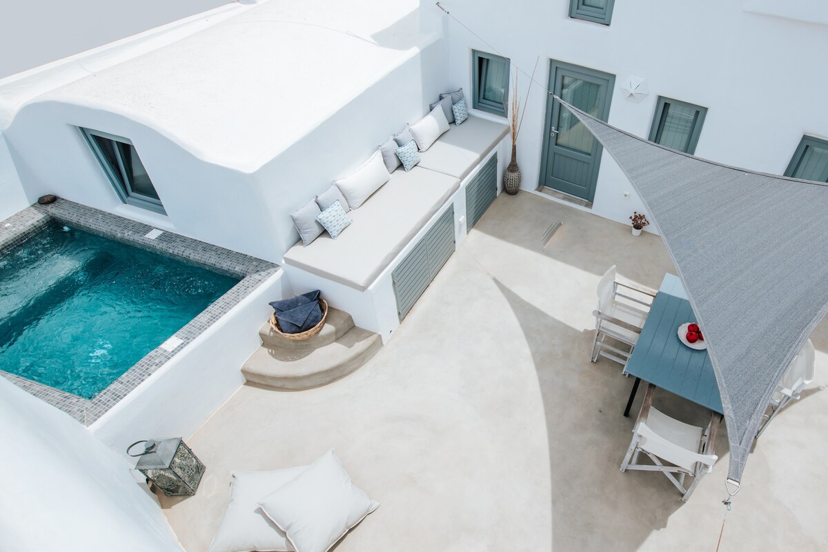 An outdoor area features a private jacuzzi tub beside a comfortable seating space with plush cushions. A dining table is positioned nearby, complemented by a large shade sail that provides protection from the sun. Well-maintained whitewashed walls encapsulate the inviting setting.