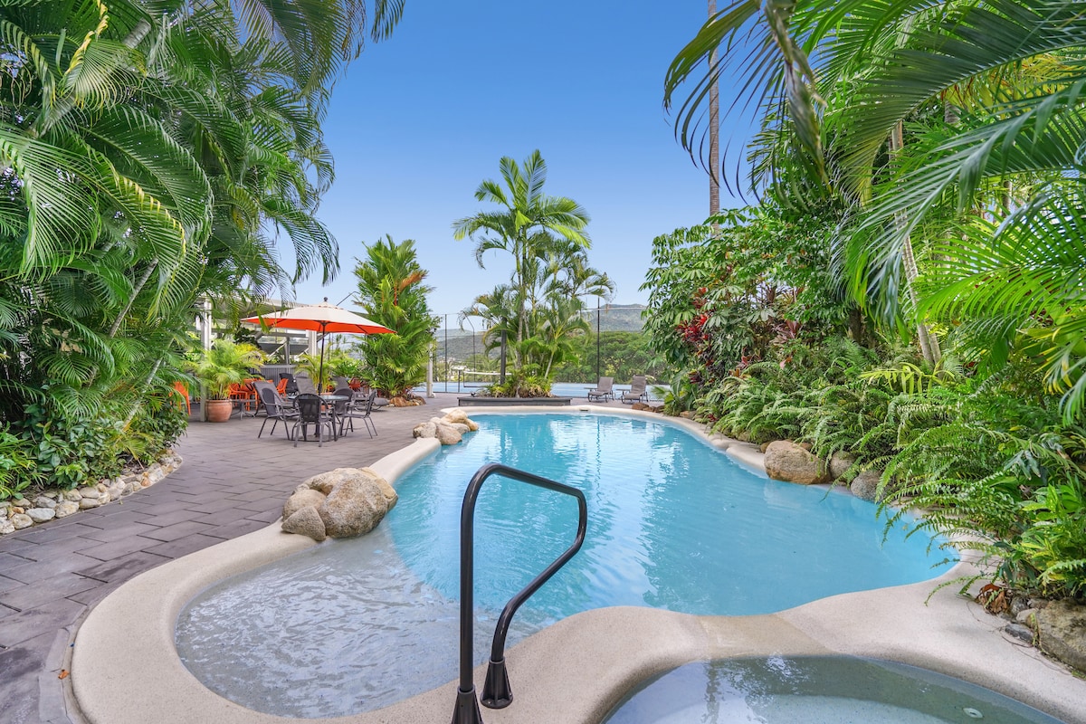 A resort-style pool is surrounded by lush tropical plants, enhancing the serene atmosphere. The water appears inviting and blue, with smooth curves and natural stone accents. Nearby, a dining set with chairs and an umbrella provides a shaded area for relaxation.
