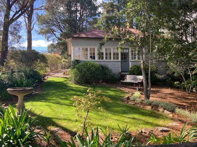 The cottage is framed by a lush garden, featuring a well-kept lawn and mature trees. The exterior showcases large windows and a welcoming entrance. A stone path leads to the dwelling, while a birdbath adds a touch of charm to the tranquil outdoor setting.