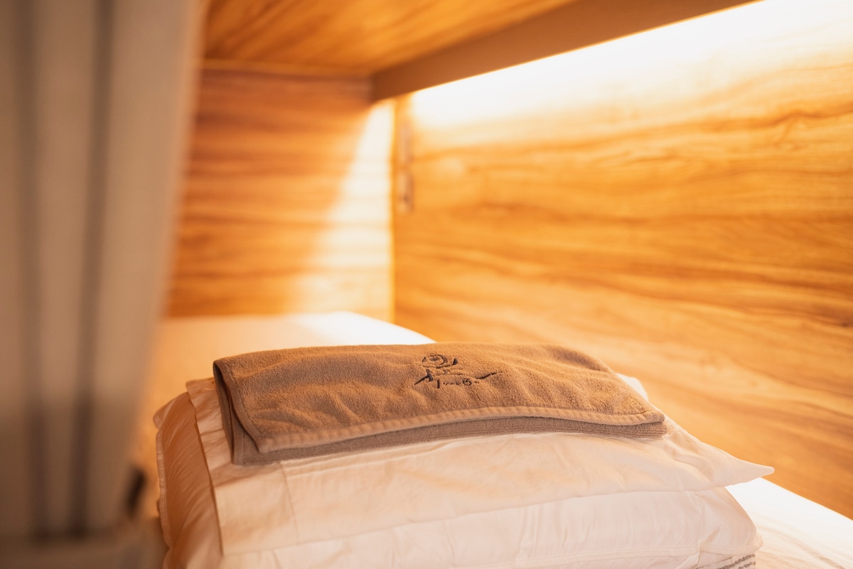 A cozy capsule bed is shown, featuring a neatly arranged stack of pillows and a soft blanket placed on top. Warm lighting from adjacent wood paneling creates a serene setting, enhancing the tranquil atmosphere of the private space.