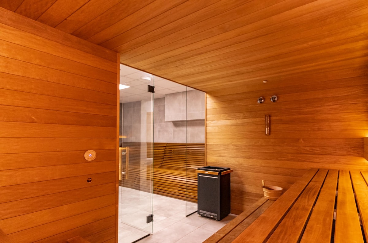 A modern sauna is enclosed with wooden paneling, creating a warm atmosphere. Wooden benches line the interior, while a glass wall provides visibility into the space. Essential amenities include a temperature control panel and a waste bin situated nearby.
