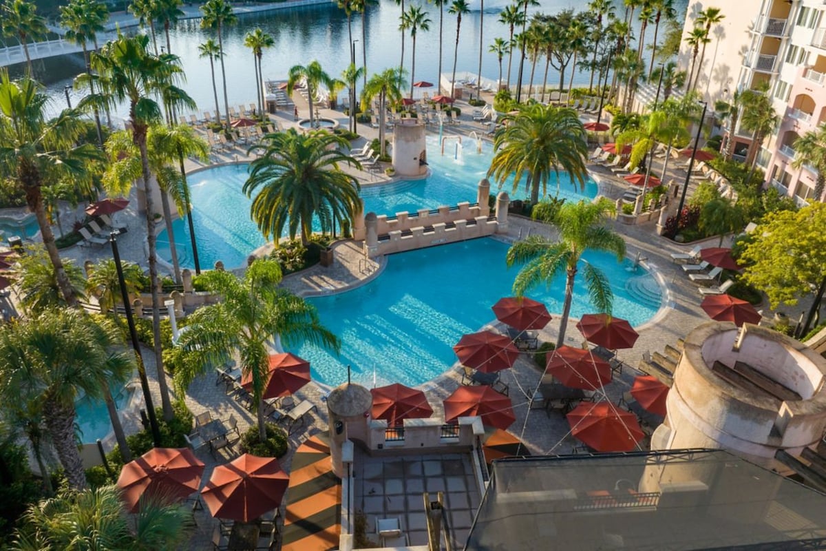 A spacious outdoor swimming pool area is surrounded by tropical palm trees and colorful umbrellas. Several loungers are positioned near the water, creating a relaxing atmosphere. A serene lake reflects the surroundings, providing a scenic backdrop to the resort amenities.