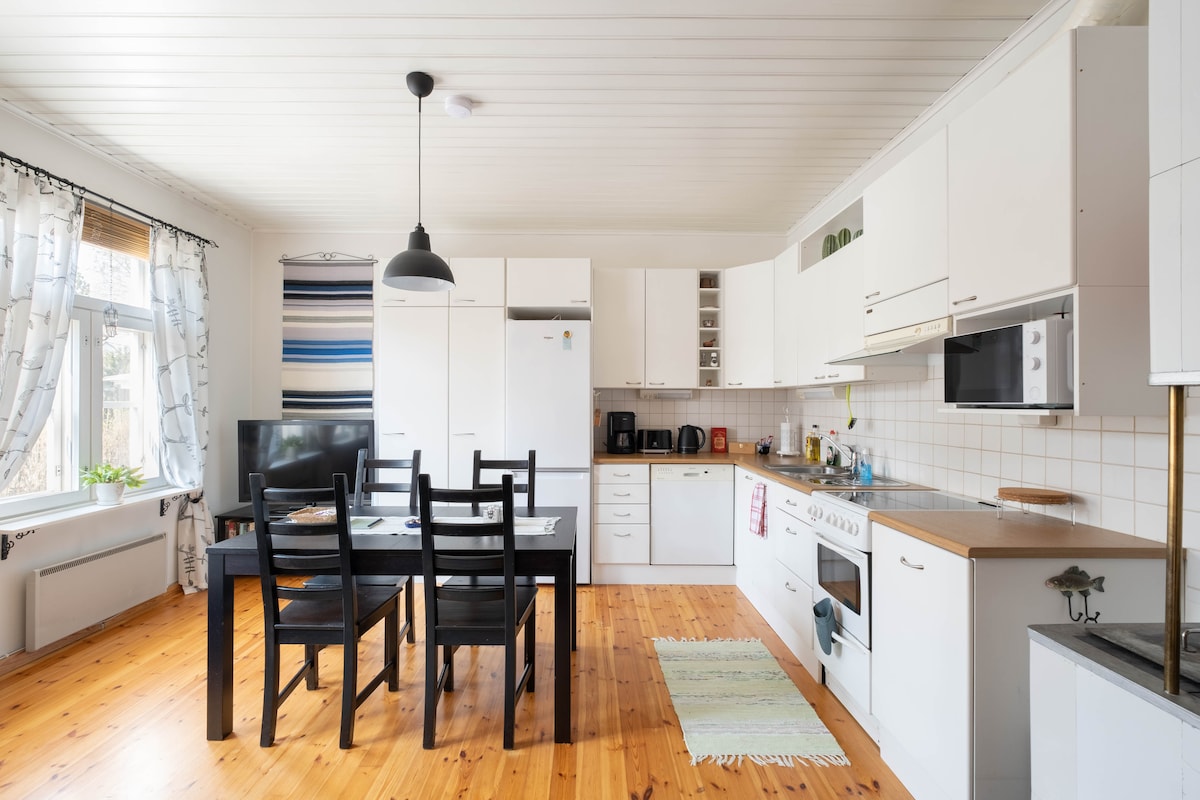 A spacious kitchen area features white cabinetry and a wooden dining table with black chairs. A television is positioned nearby, and various kitchen appliances, including a stove, microwave, and refrigerator, are neatly arranged. Large windows allow natural light to brighten the space.