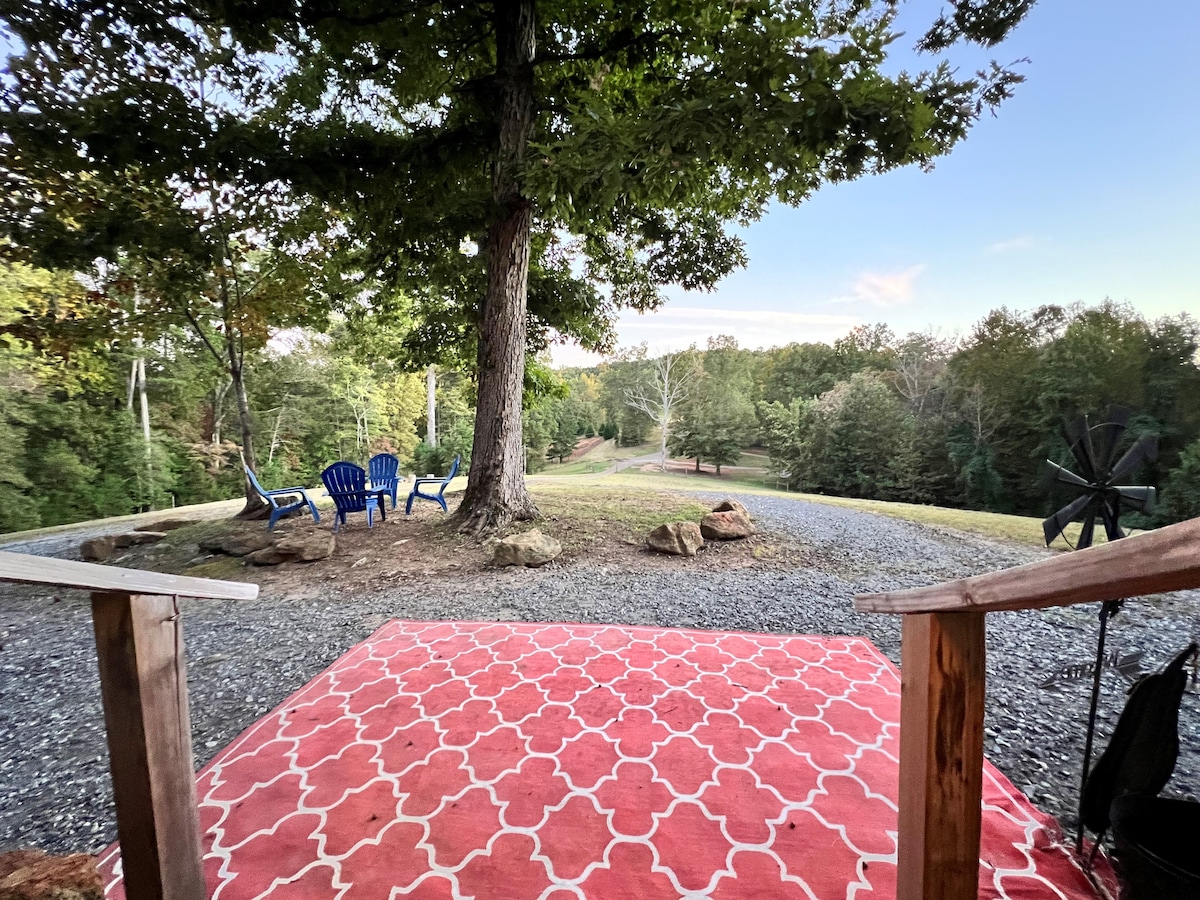 An outdoor seating area is set beneath a large tree, featuring two blue chairs facing a gravel pathway leading through a scenic landscape. A patterned rug adds comfort, and a wind spinner is visible nearby, contributing to the tranquil atmosphere.