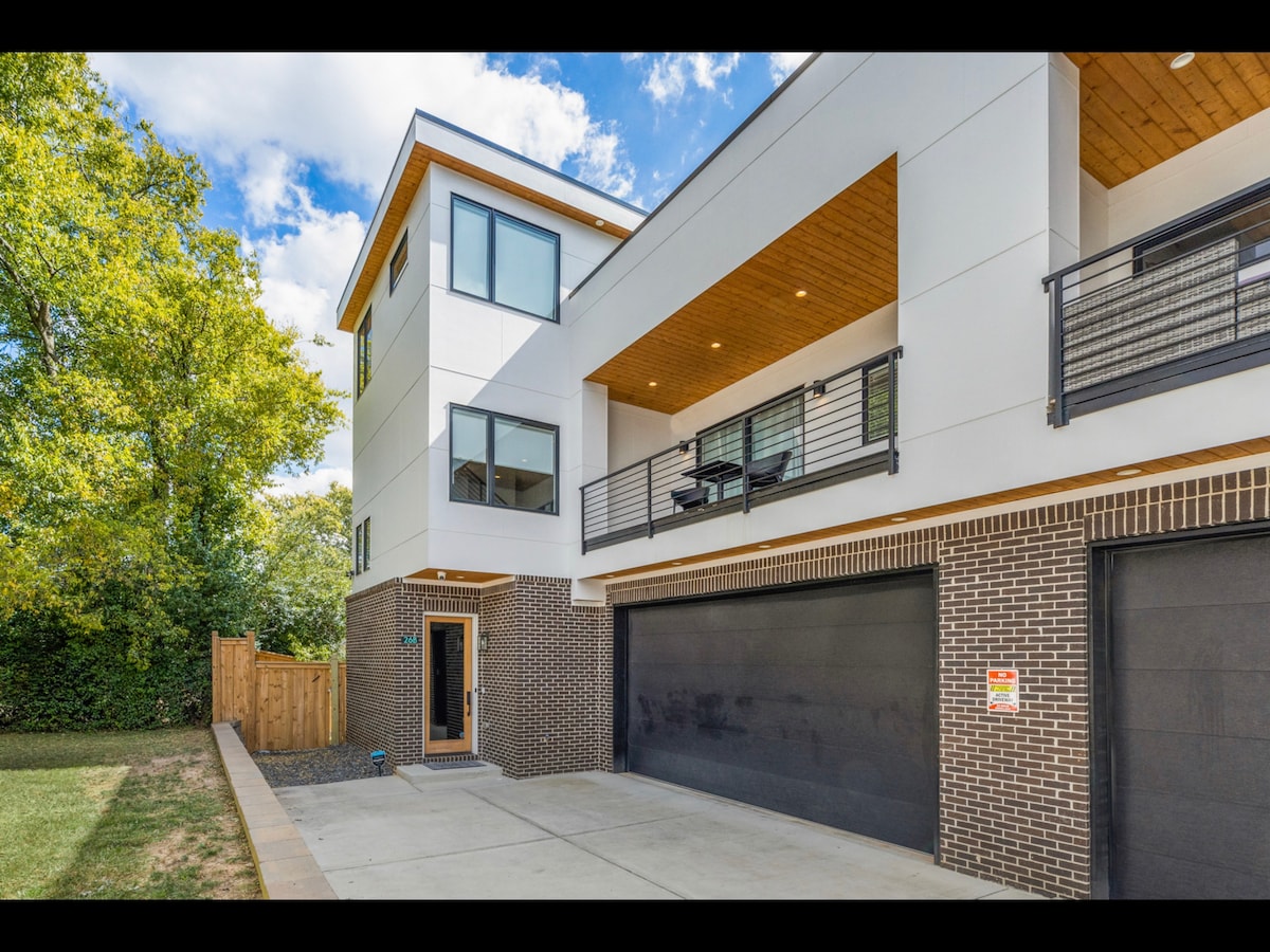 The modern exterior of the home features a combination of white siding and dark brick. A covered balcony extends from the second floor, overlooking the front area. The two-car garage is visible, set against a backdrop of trees and a serene yard.