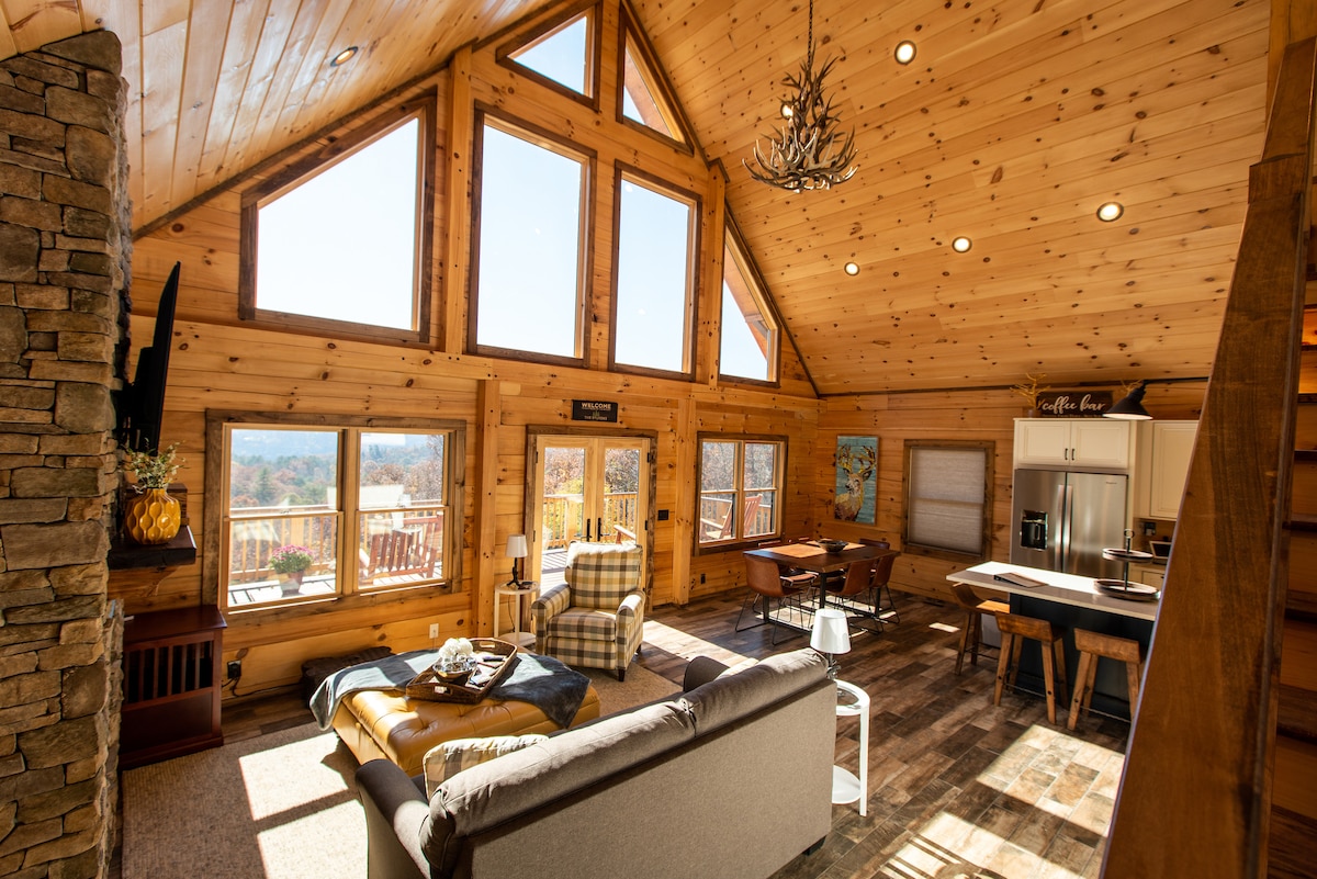A spacious living area features large triangular windows that allow natural light to fill the space, offering views of the outdoors. Wooden walls and ceilings create a rustic charm. A cozy sofa is paired with an armchair and a dining table, complementing the aesthetic of the room.