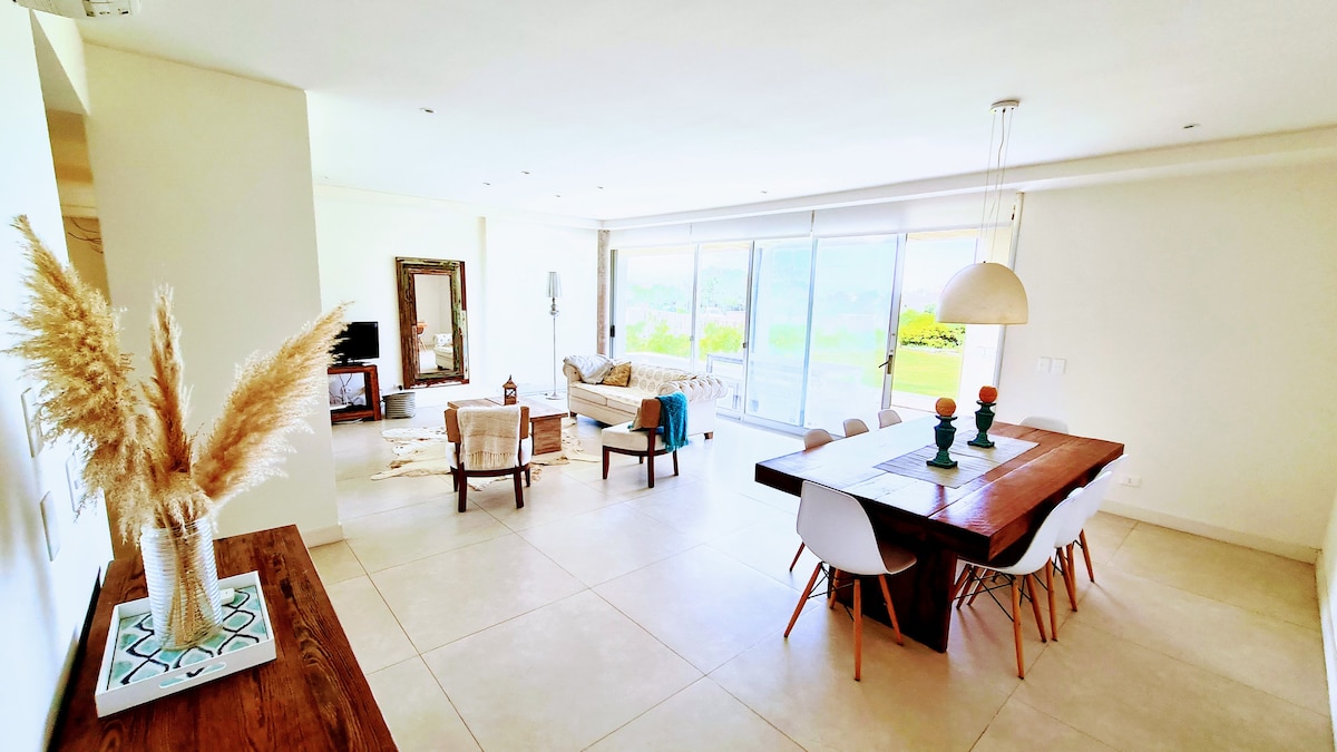 An open living and dining area is presented, featuring a large dining table with white chairs. Soft natural light enters through expansive glass doors, illuminating a comfortable seating arrangement with a neutral color palette. Decorative elements include a plant and a mirror on the wall.