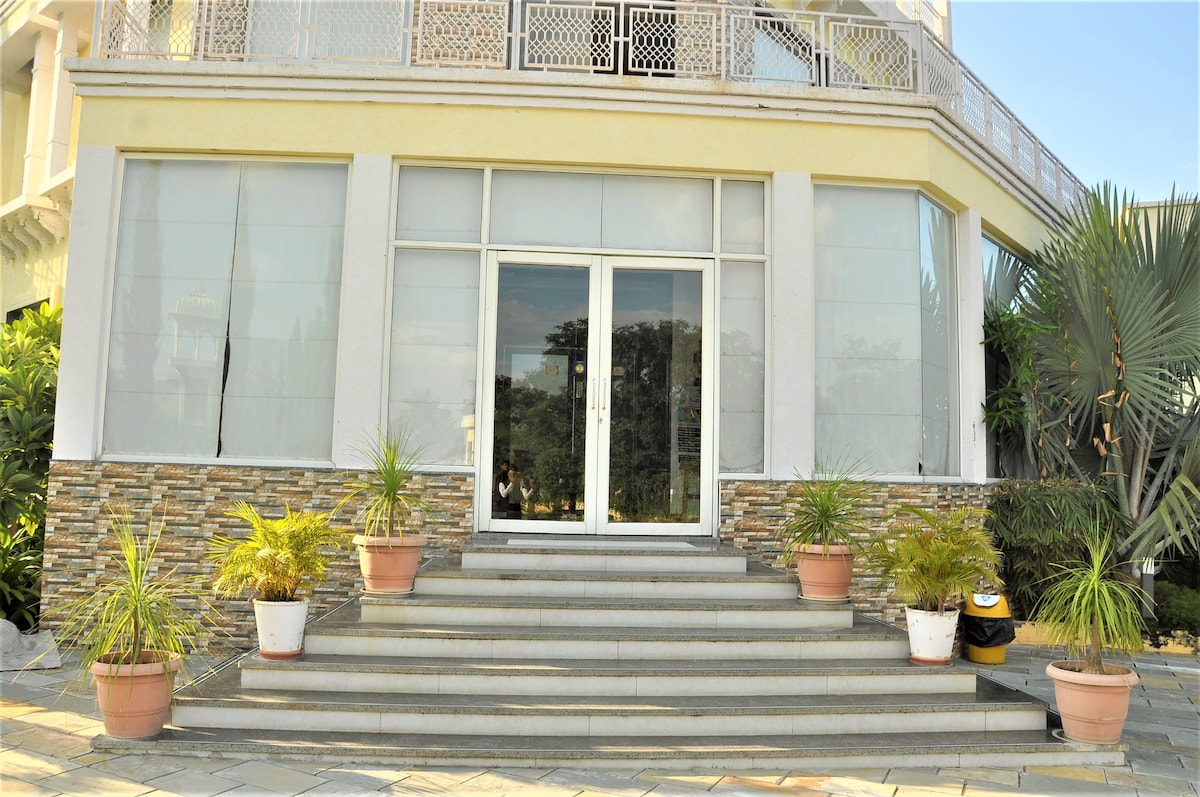 The entrance of the property is showcased, featuring a series of stairs leading up to large glass doors. Potted plants are arranged on either side of the steps, adding greenery to the modern façade. Natural light is evident, enhancing the welcoming appearance.