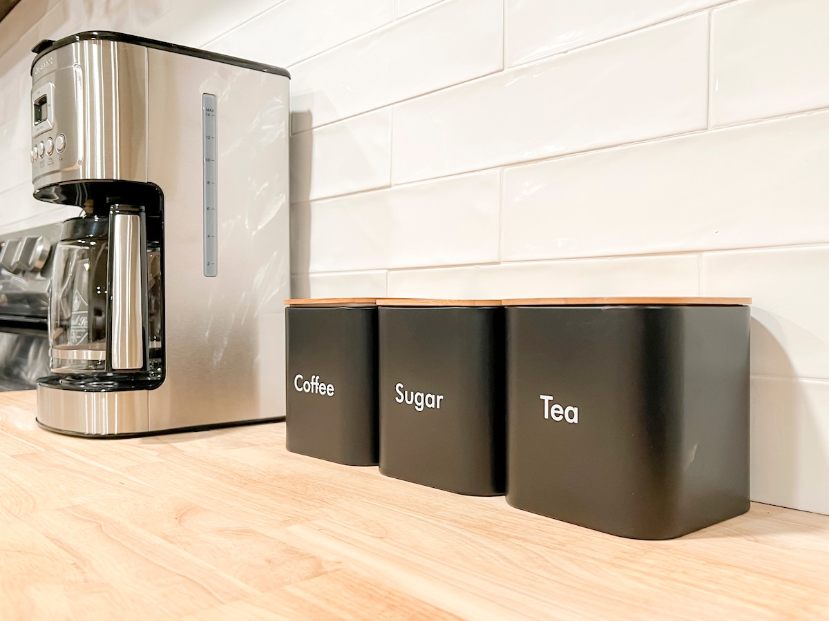 A modern kitchen countertop displays a coffee maker next to three black storage containers labeled 'Coffee,' 'Sugar,' and 'Tea.' The sleek containers are complemented by a light-colored wooden surface and white subway tile backsplash.