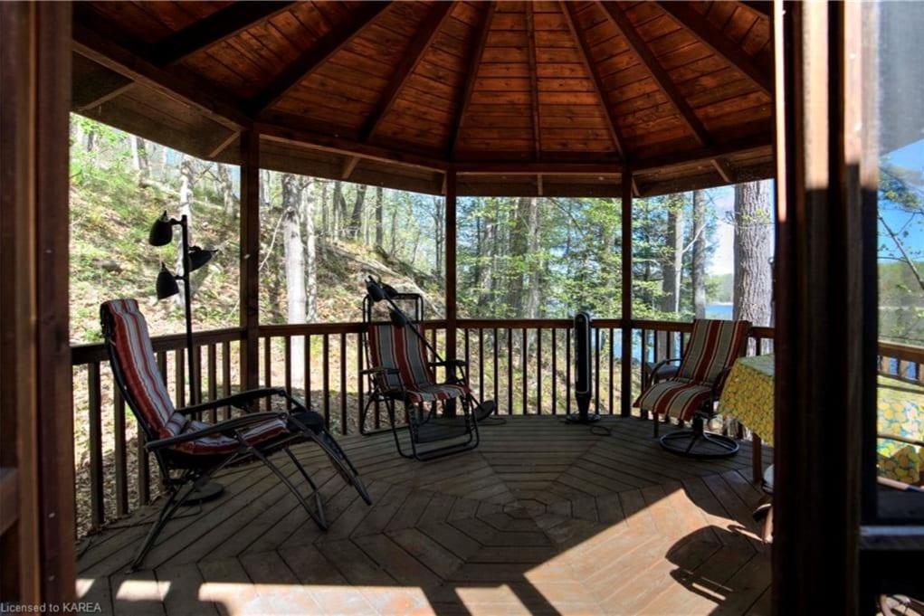 A screened gazebo features wooden flooring and an octagonal shape, with multiple seating areas including two reclining chairs and a small table draped with a yellow cloth. Panoramic views of the surrounding forest and nearby lake are visible through the mesh screens.