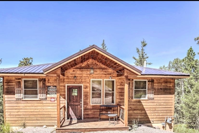 The cabin exterior is characterized by warm wooden siding and a modern metal roof. Large windows allow plenty of natural light, and a welcoming front porch features a small seating area. The surrounding landscape consists of tall pines under a clear blue sky.