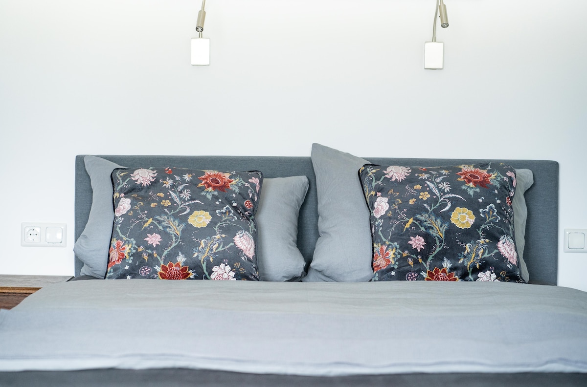A spacious bed is presented with light gray bedding and decorative pillows featuring floral patterns. The headboard, in a muted tone, complements the overall calming color scheme. Wall-mounted lights are positioned above the bed, providing a clean and modern touch to the sleeping area.