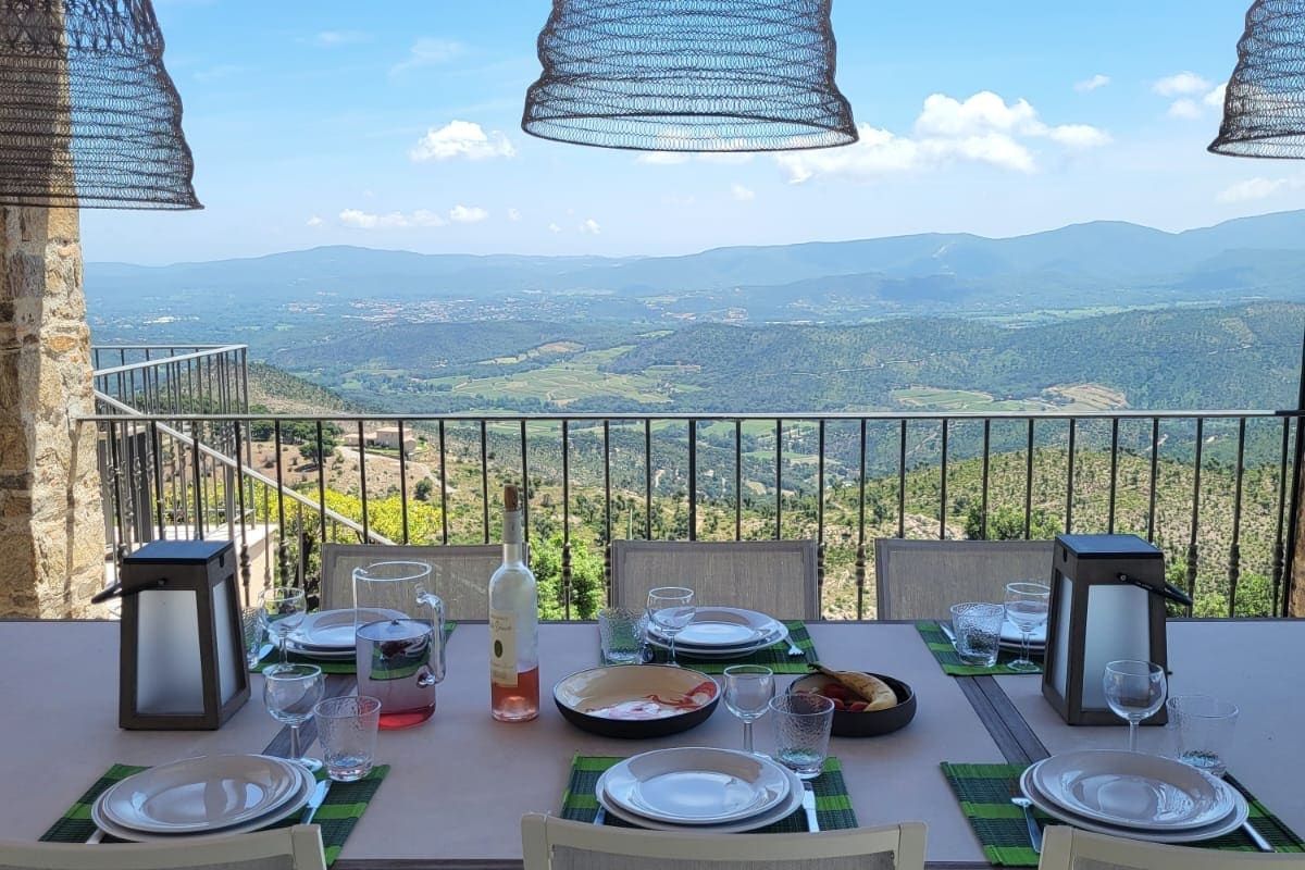 A dining table is set outdoors with scenic views of the surrounding hills and valleys. The table features glassware, plates, and utensils, accompanied by lanterns. The shade from woven light fixtures enhances the serene ambiance, inviting guests to enjoy meals in a picturesque setting.