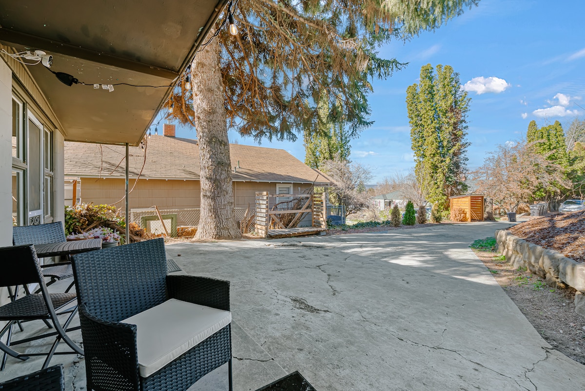 An outdoor patio area features black wicker seating with cream cushions, situated on a concrete surface. A gently sloping yard with trees is visible in the background, alongside a private driveway leading to the home, creating a peaceful and spacious environment.