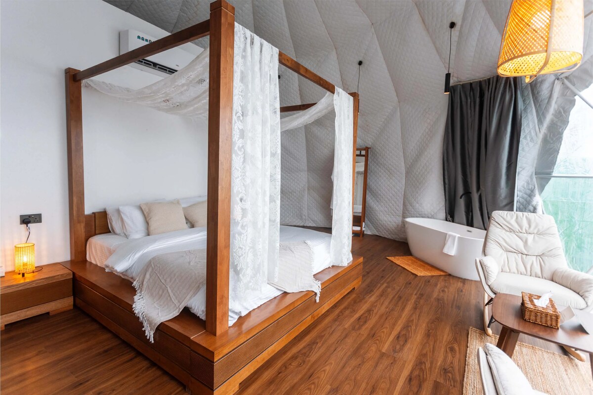 A spacious double bed with a wooden frame is draped in soft, delicate fabrics, complemented by bedside lamps. A cozy seating area includes an elegant chair and a wooden table. A freestanding bathtub offers a serene view through large windows, enhancing the tranquil ambiance.