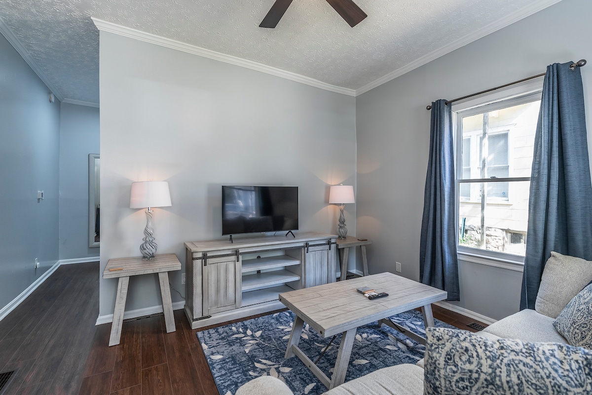 A cozy living area displays a comfortable sofa, complemented by a light gray wall. A coffee table sits centrally, with a television mounted on a light wood stand. Natural light filters through two large windows, dressed with blue curtains.