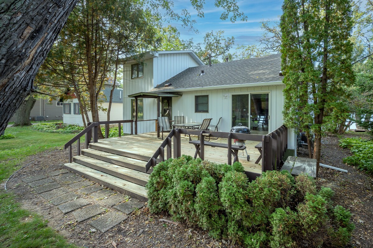 A private deck area is shown with wooden steps leading up to it. Simple outdoor furniture is arranged for seating, accompanied by a BBQ grill. Lush greenery surrounds the space, providing a natural backdrop to the modest, two-story retreat.
