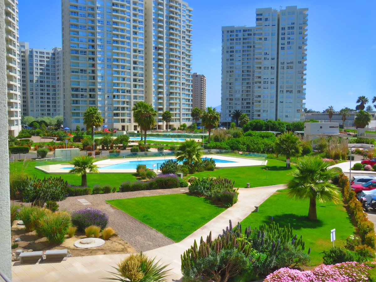 An outdoor area featuring a well-maintained garden with various plants and palm trees. A swimming pool is visible at the center, surrounded by neatly manicured green spaces. Towering residential buildings line the background under a clear blue sky.