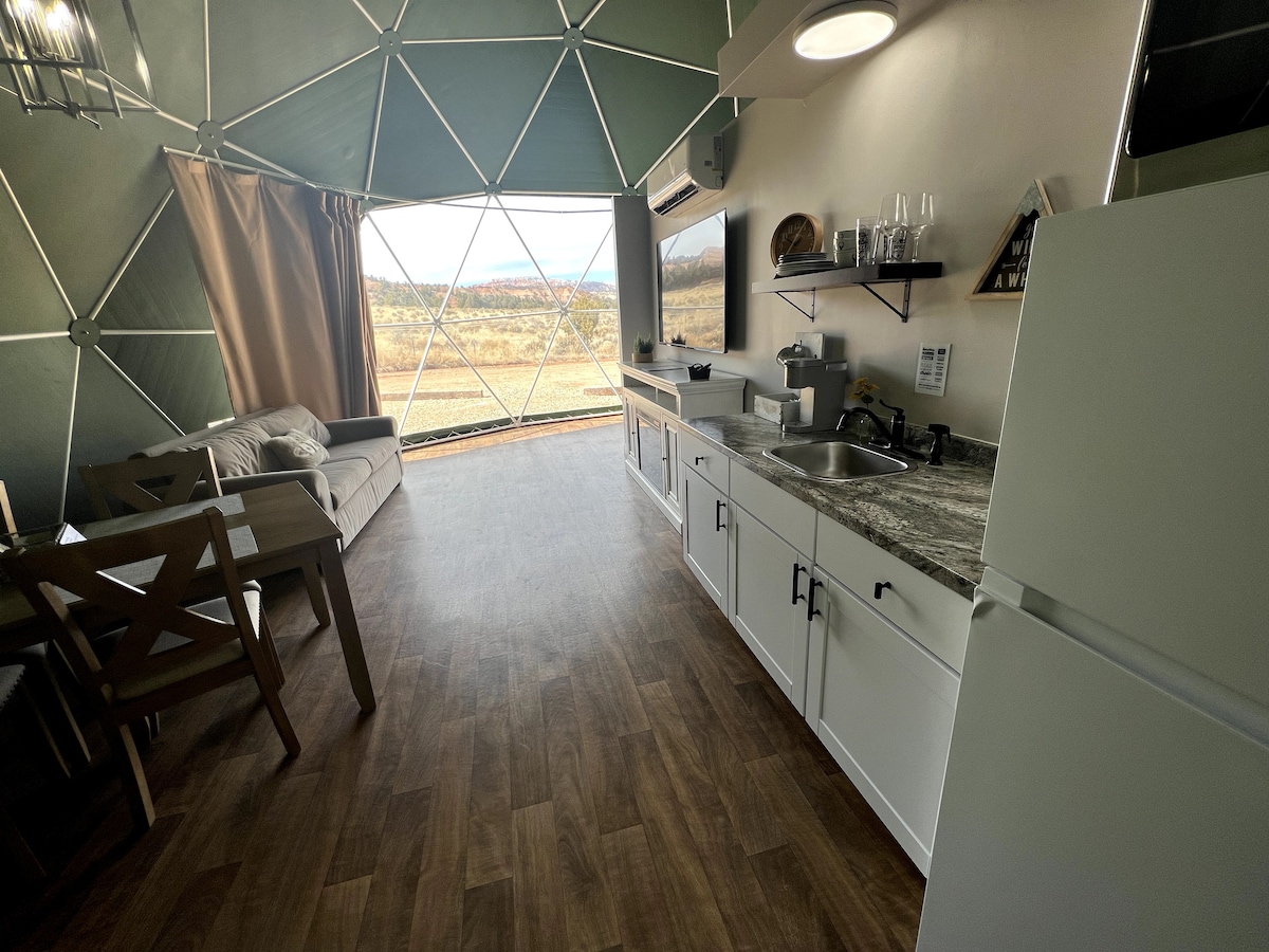 An interior view of a glamping dome features a modern kitchen area with a stone countertop and appliances. A cozy seating arrangement is positioned next to a large window, providing views of the surrounding landscape. Natural light fills the space through the dome's signature design.