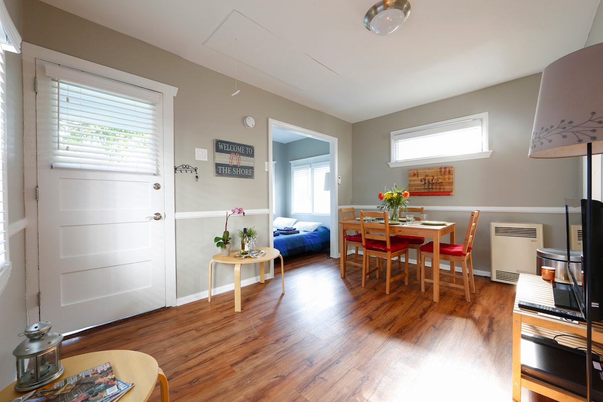 A bright and airy living space includes a white entry door and a cozy seating area with a round table and four chairs. Natural light bathes the laminate flooring, with a glimpse of the blue bedroom visible in the background, enhancing the bungalow's inviting charm.