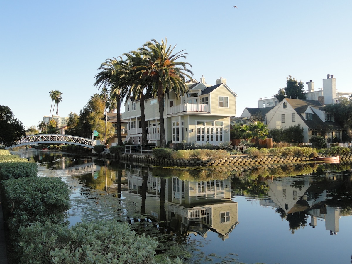 A scenic canal lined with lush greenery features a charming two-story house with a balcony overlooking the water. Palm trees flank the property, reflecting beautifully in the calm surface of the canal, creating a serene and inviting atmosphere.
