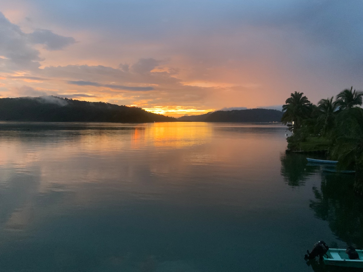 A vibrant sunset is reflected on the calm waters of the bay, surrounded by lush tropical hills. Silhouettes of palm trees line the shoreline, and the sky is painted with hues of orange, pink, and purple as the day transitions to evening.
