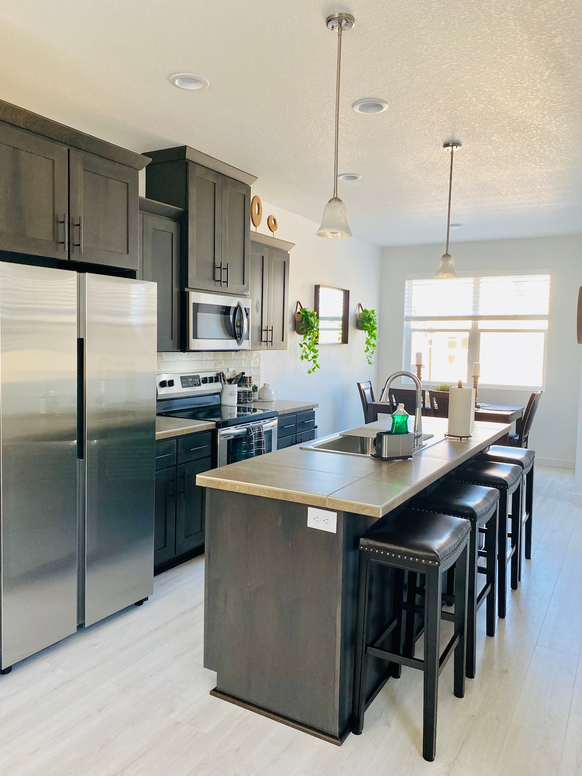 A modern kitchen features sleek black cabinetry and stainless steel appliances. A spacious island with seating for four complements the cooking area. Light-colored countertops enhance the bright atmosphere, while natural light filters through a nearby window. Decorative plants add a touch of greenery.