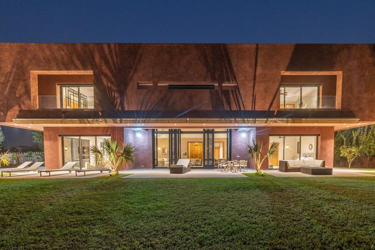 The contemporary villa features a warm rust hue and expansive glass doors, integrating indoor and outdoor living. Soft lighting highlights the manicured lawn and surrounding palm trees, while outdoor seating areas are arranged for social gatherings under the evening skies.