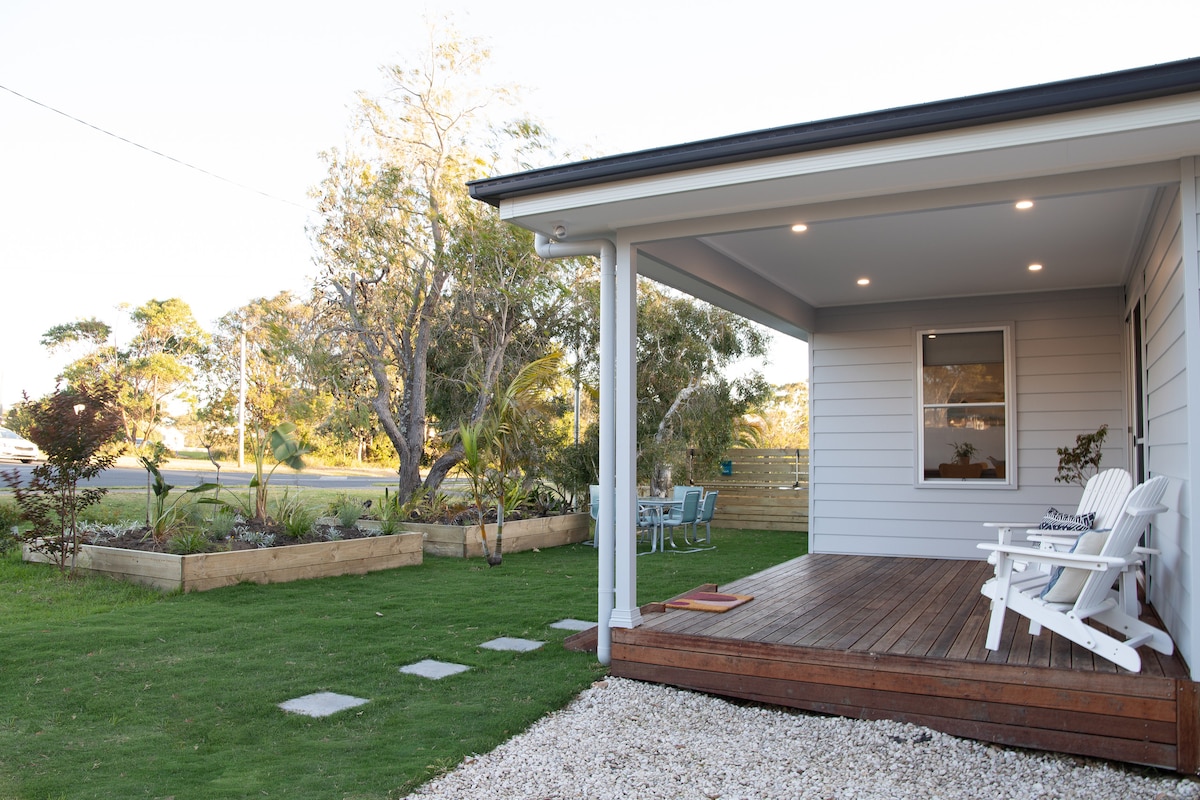 A welcoming outdoor area features a wooden deck with white rocking chairs, surrounded by a well-maintained lawn. Native plants and shrubs are visible in garden beds, while an outdoor dining set is positioned nearby, offering a relaxing space for enjoyment.