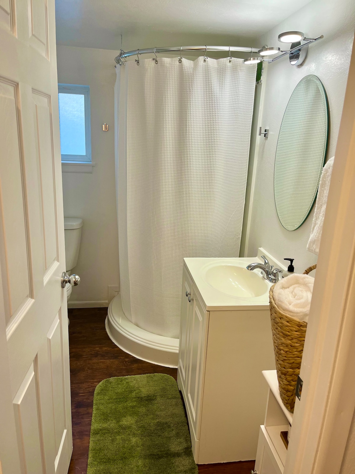 A compact bathroom features a round shower stall with a white curtain and a small vanity with a sink. A wall-mounted mirror reflects the space. Neutral-colored walls complement a green bath mat and woven basket that holds towels. A toilet is positioned in a separate alcove.