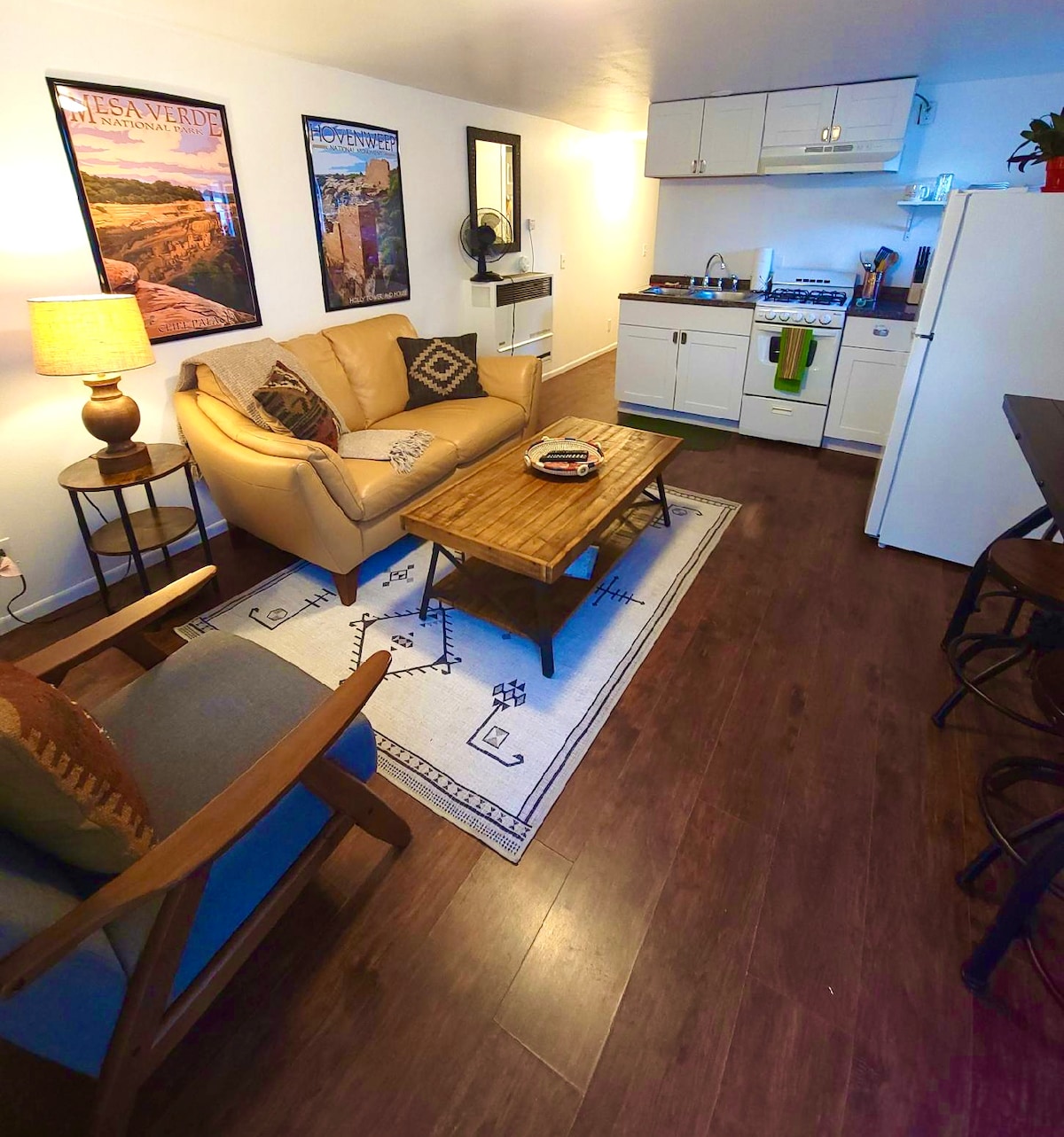 A cozy living area is showcased, featuring a beige sofa and a wooden coffee table. Framed artwork adorns the walls, complementing the warm lighting from a bedside lamp. An efficient kitchen area is visible, equipped with white cabinetry and essential appliances.