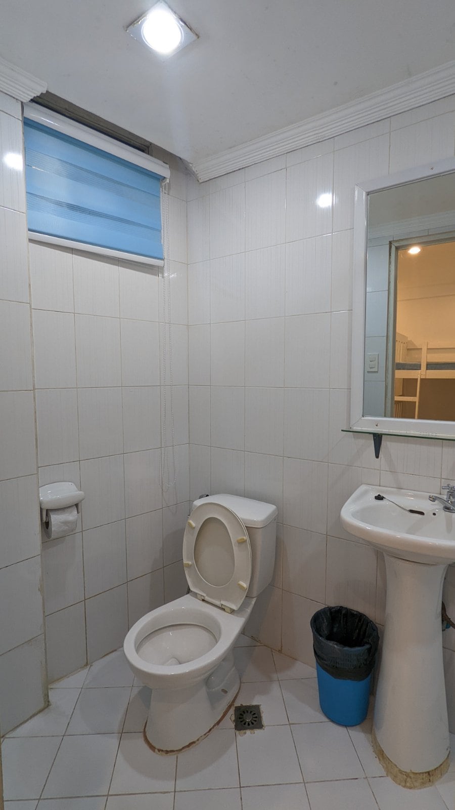 The private bathroom includes a white toilet and a pedestal sink positioned next to a wall-mounted mirror. A blue trash bin is visible on the floor, and a window covered with light blue blinds allows natural light to enter the space.