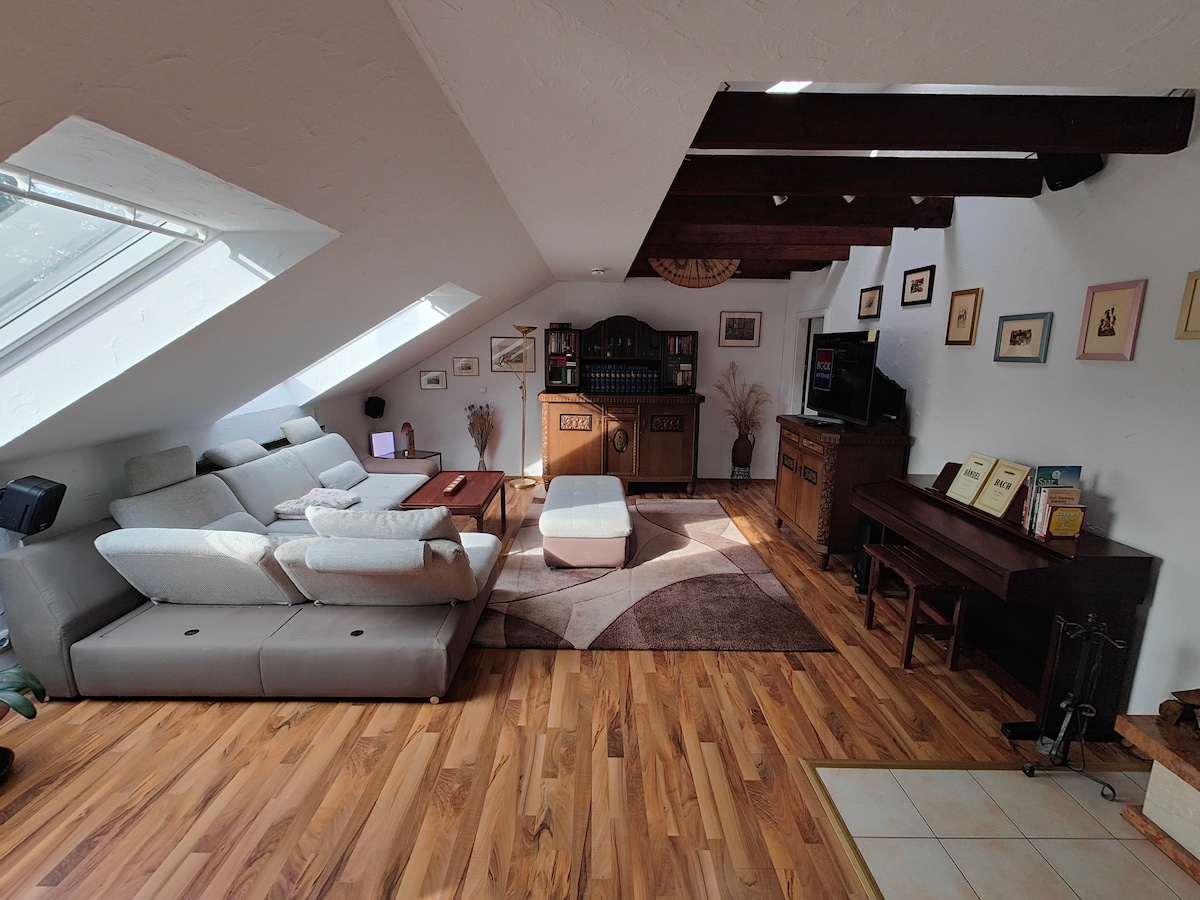 A spacious living area features a large sectional sofa positioned around a coffee table, with wooden flooring throughout. Natural light is let in through angled skylights. A piano sits in one corner, complemented by shelves holding books and decor on the walls.