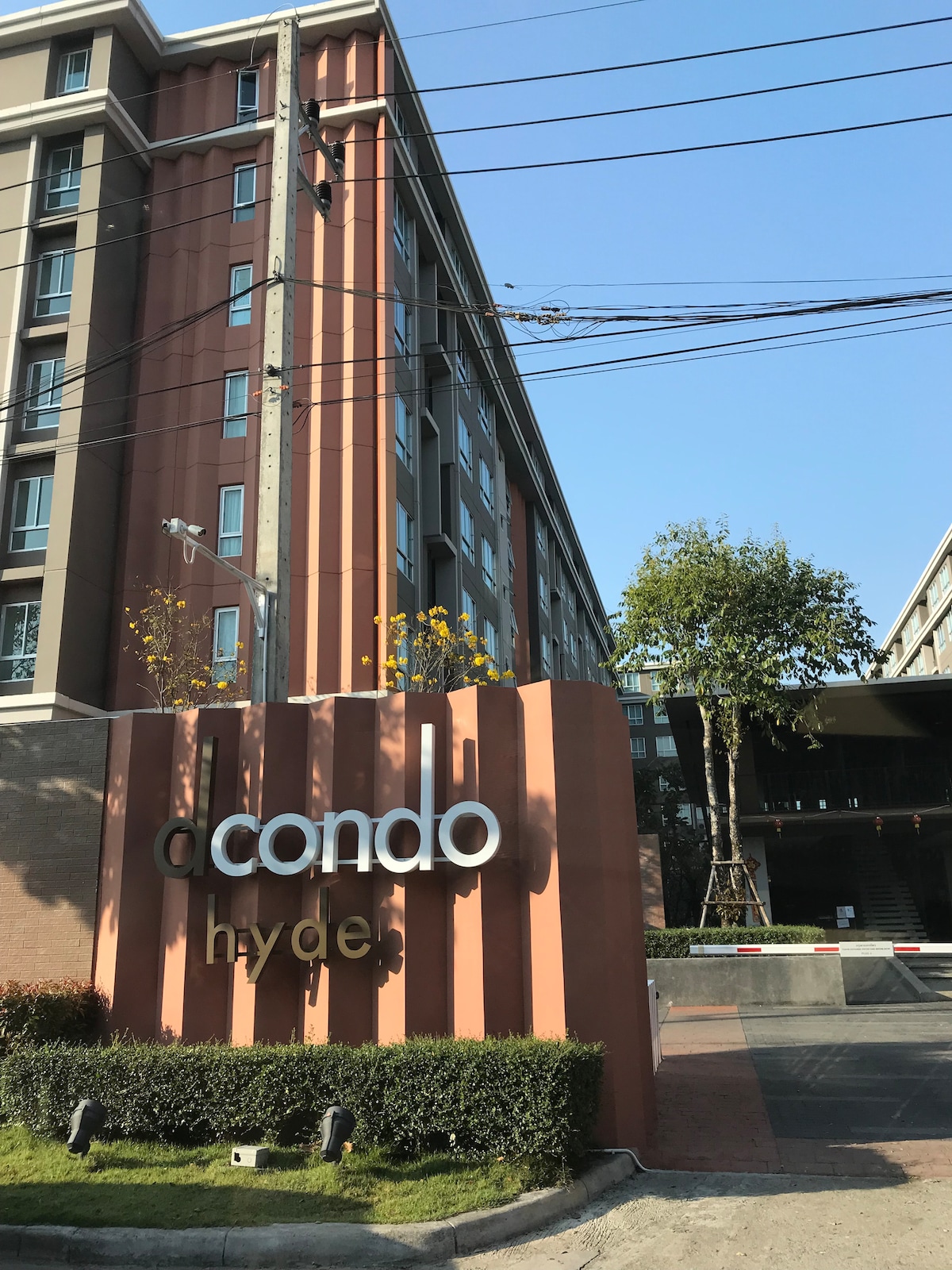 The exterior of a modern condominium building is showcased, featuring clean lines and a contemporary design. A prominent sign displays the name 'a condo hyde,' surrounded by greenery and pedestrian pathways. A clear blue sky enhances the urban environment.