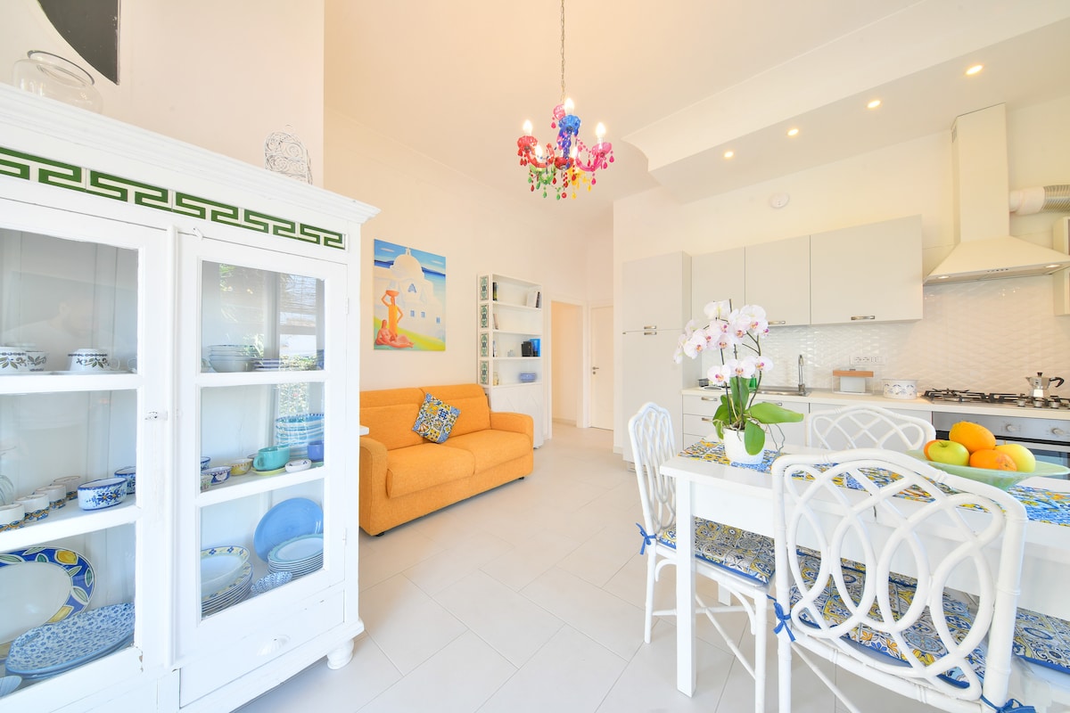 A bright living space is filled with natural light, featuring an orange sofa and a white dining table set with colorful tableware. A glass cabinet displays decorative dishes, while a pendant chandelier adds a touch of color to the ceiling. Fresh fruit is arranged on the table.