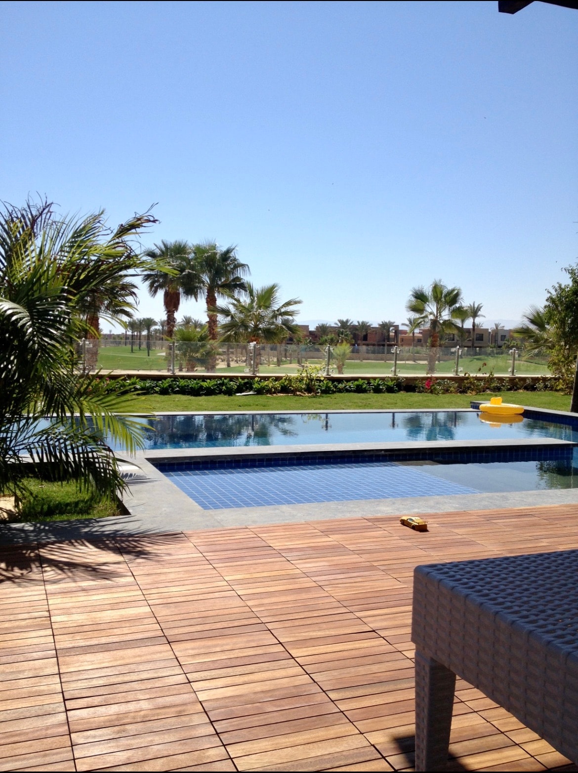 An outdoor view features a modern infinite pool bordered by wooden decking. Lush tropical plants and palm trees create a vibrant setting, overlooking a vast green golf course. Clear blue skies enhance the tranquil ambiance of the space.