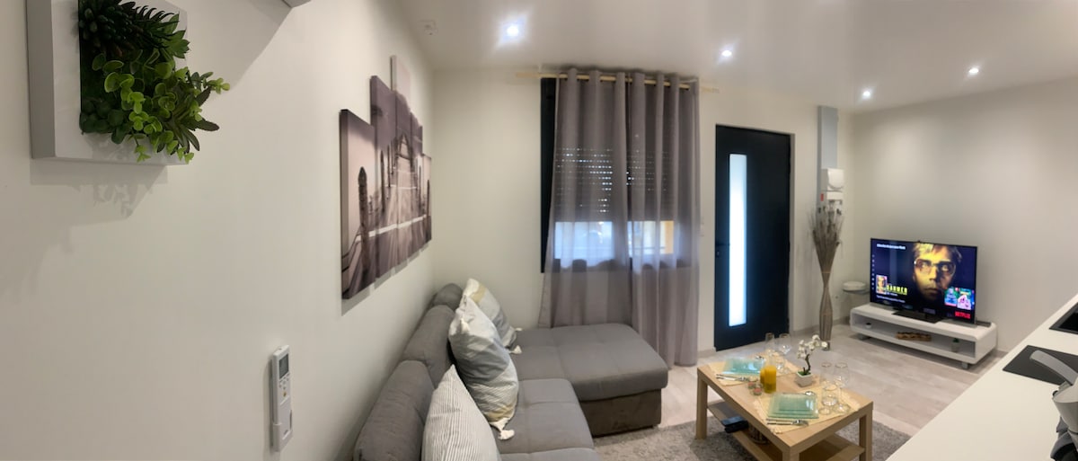 A cozy living area features a gray sofa with decorative cushions and a coffee table. A television is positioned on a white stand, and soft lighting enhances the space. Vertical curtains frame a window, contributing to a bright yet inviting atmosphere.