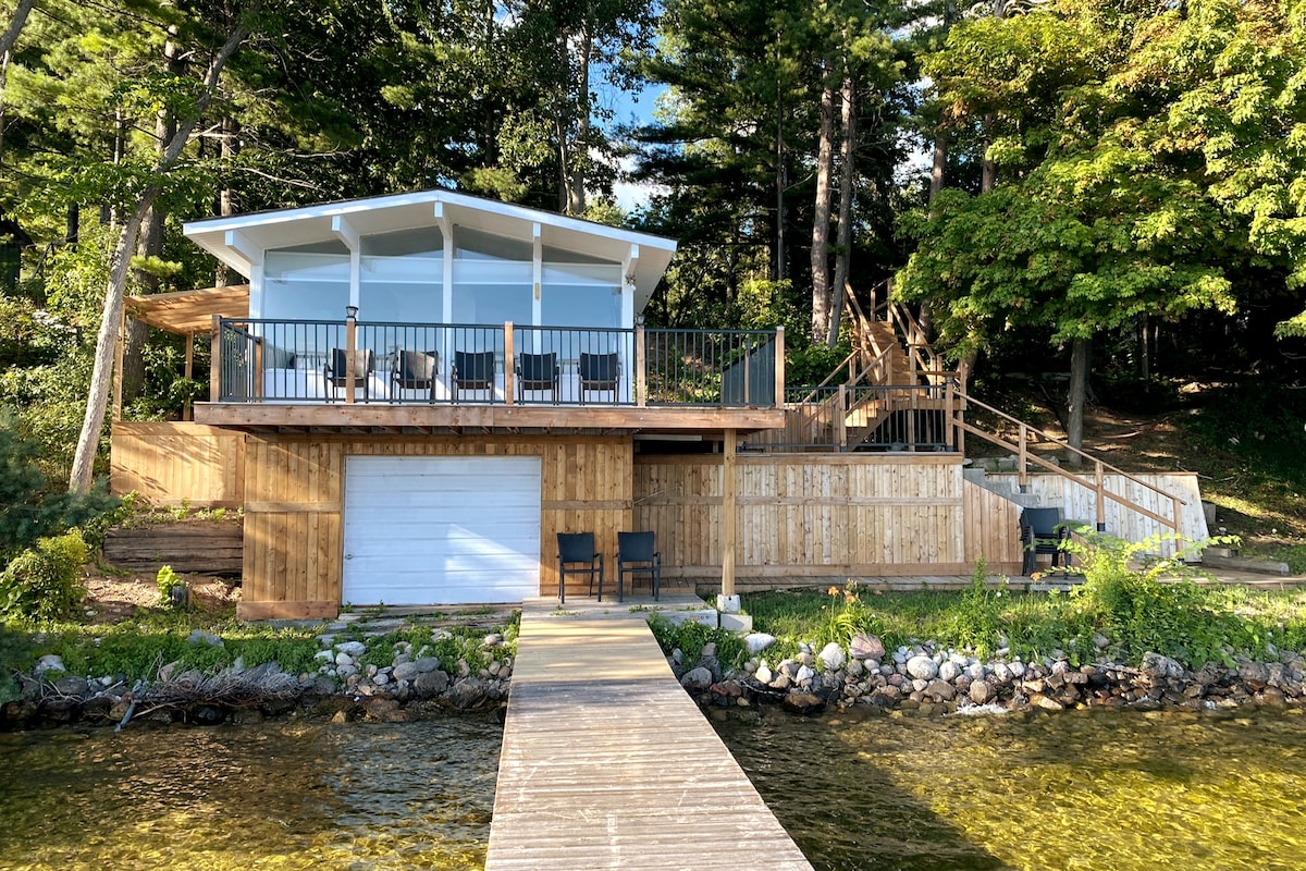 A charming boathouse is depicted with a wooden exterior surrounded by greenery. A spacious deck offers seating and overlooks the lake. A private dock extends into the water, providing access. Steps lead from the deck to the lush landscape and entrance.