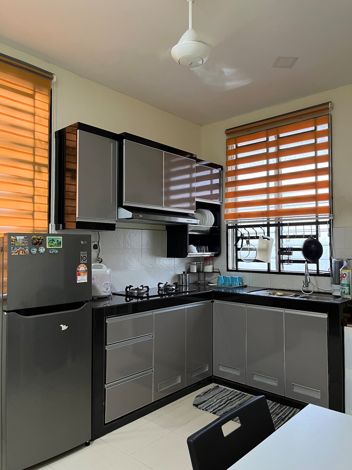 A modern kitchen is equipped with gray cabinets and black countertops. A refrigerator and gas stove are prominently displayed. Sunlight filters through orange striped blinds, creating a warm ambiance. A small dining table is positioned nearby, complementing the functional layout of the space.