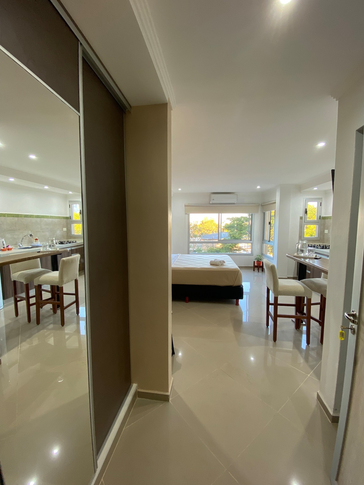 A well-lit interior showcases an open-plan layout connecting the kitchen and living area. Mirrors reflect the space, while a spacious bed is visible in the background. Barstools accompany a kitchen island, and natural light streams through the large window, enhancing the room's brightness.