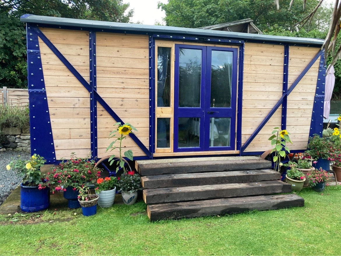 The entrance of a fully restored railway carriage is shown, featuring wooden siding and blue accents. Steps leading to the door are flanked by vibrant planters filled with blooming flowers, creating a welcoming aesthetic. Surrounding greenery enhances the outdoor environment.