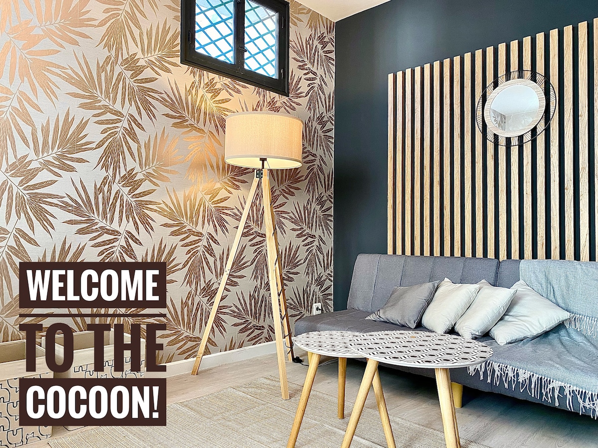 A modern living area is featured, highlighted by a decorative wall with leaf patterns and warm wooden accents. A contemporary floor lamp stands beside a grey sofa adorned with cushions. A round mirror reflects light, enhancing the space's inviting atmosphere.