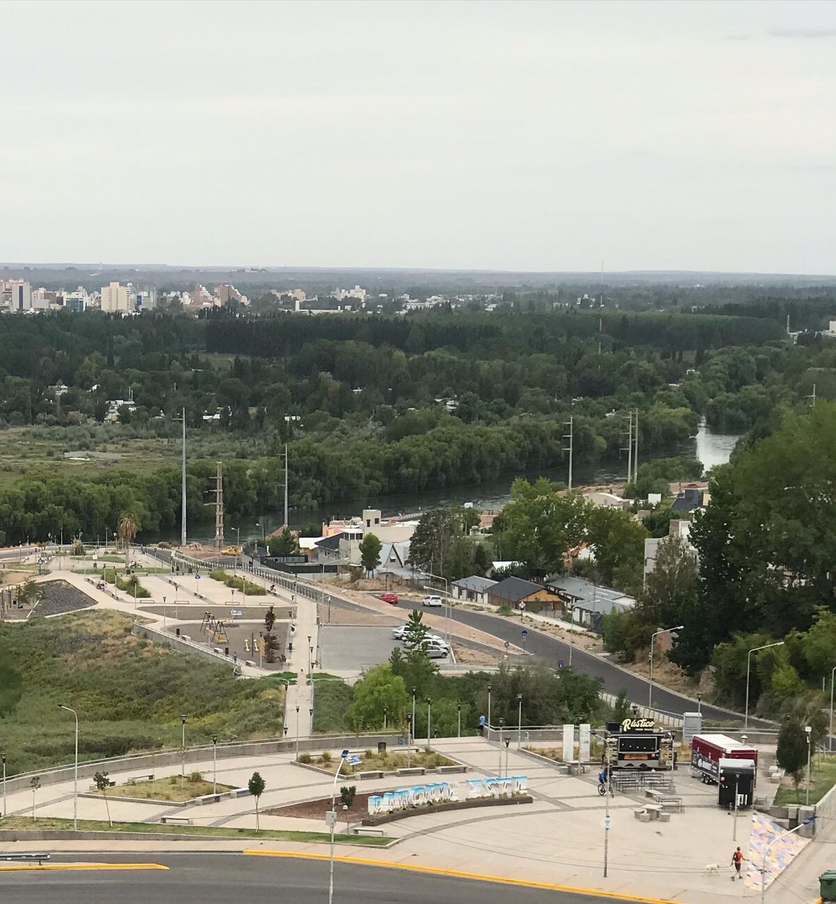 A scenic view reveals the river winding through greenery, with city buildings visible in the distance. The pathway along the riverbank is well-defined, featuring trees and shrubs, creating a natural contrast with the urban landscape beyond.