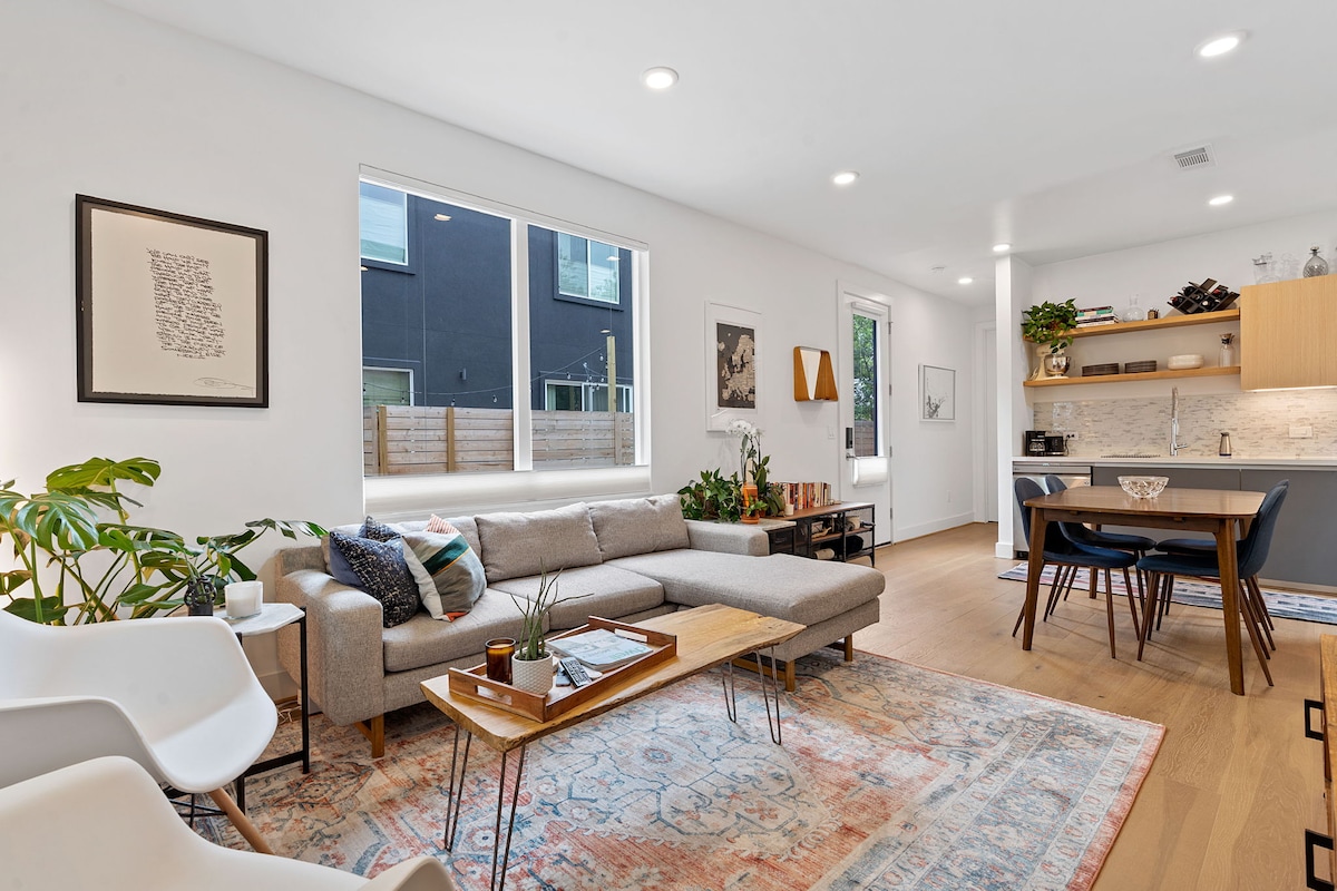 A modern living area features a light grey sectional sofa positioned near a wooden coffee table. Natural light floods in through large windows, illuminating the space. A dining table with chairs sits adjacent to a well-equipped kitchen with wooden cabinetry and various decorative plants enhancing the environment.