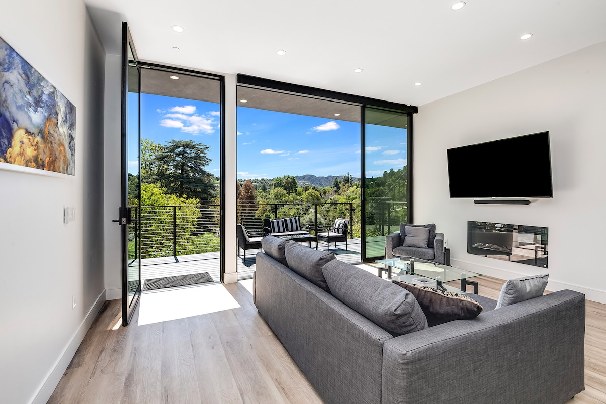 A modern living area is highlighted by large glass doors that open to a balcony with hillside views. Comfortable seating is arranged on a sleek sofa, with a clear coffee table in front. Natural light floods the space, complemented by a mounted TV on the wall.