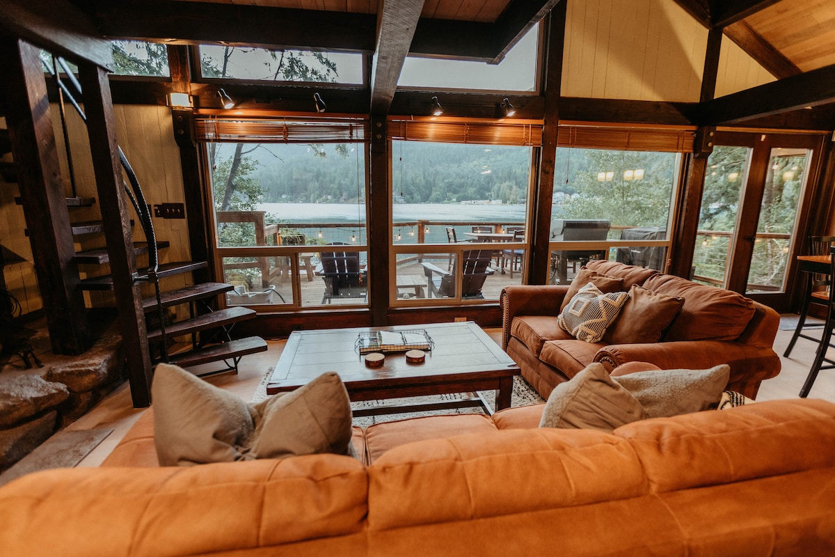 A cozy living area features comfortable sofas facing a wooden coffee table. Floor-to-ceiling windows provide a view of the lake and surrounding trees. A spiral staircase leads to an upper level. Soft lighting from multiple sources enhances the inviting setting.
